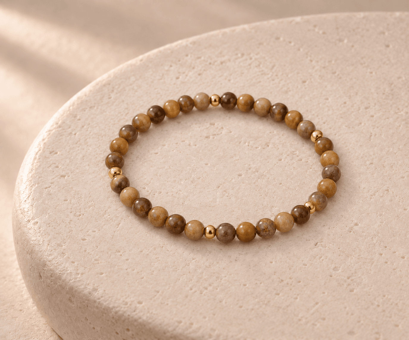 Earth bead bracelet in warm brown with small gold spacers on a rounded cream stone pedestal, without a pendant