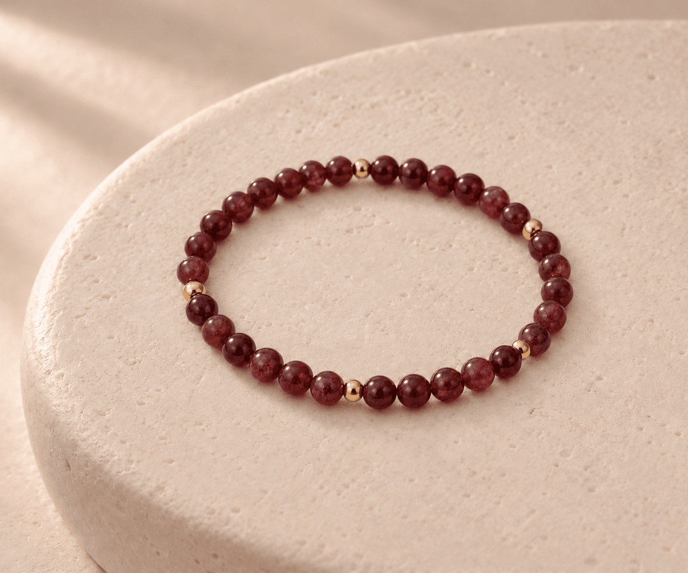 Fire bead bracelet in wine red with small gold spacers on a rounded cream stone pedestal, without a pendant