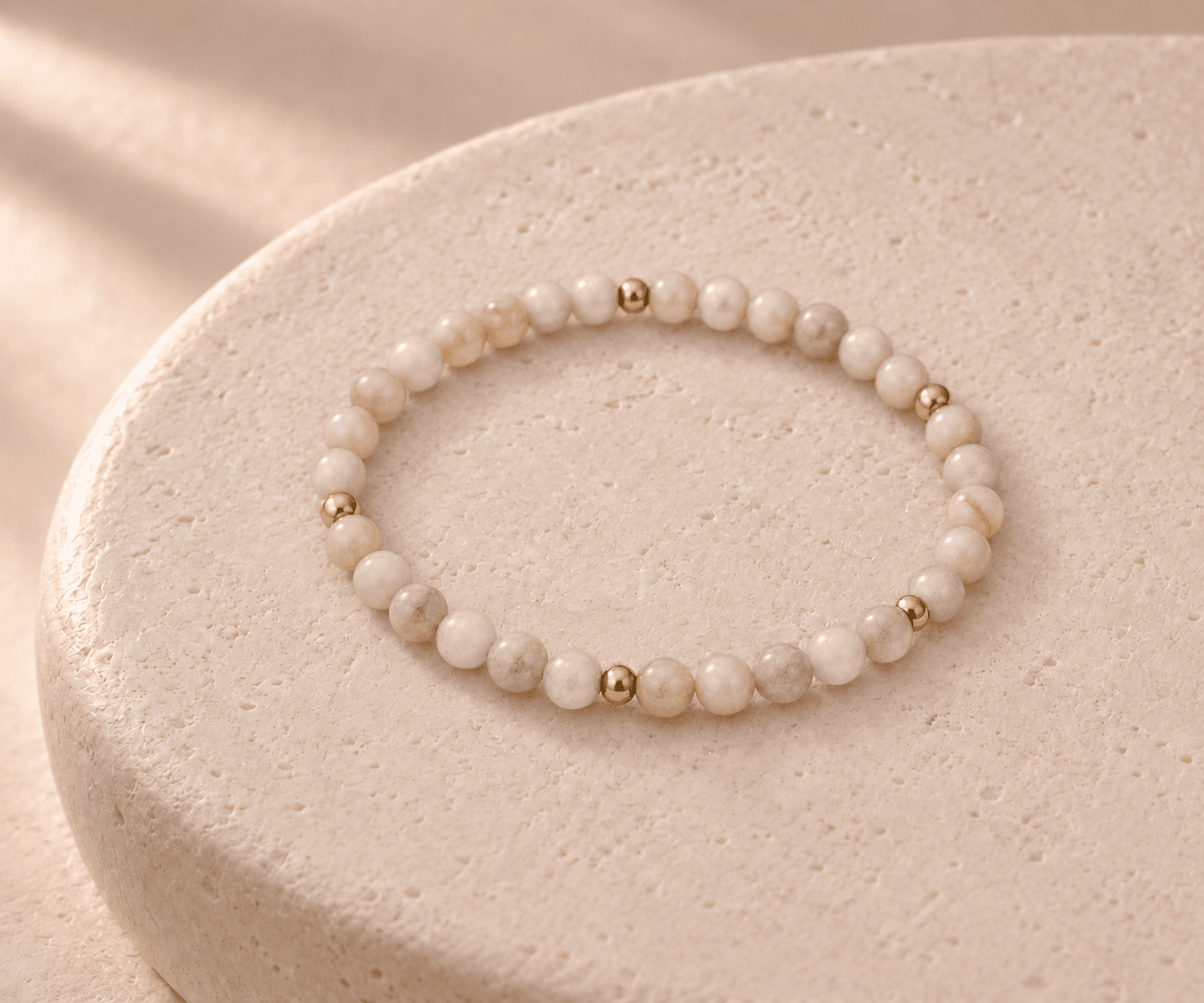 Metal bead bracelet in milky ivory with small gold spacers on a rounded cream stone pedestal, without a pendant